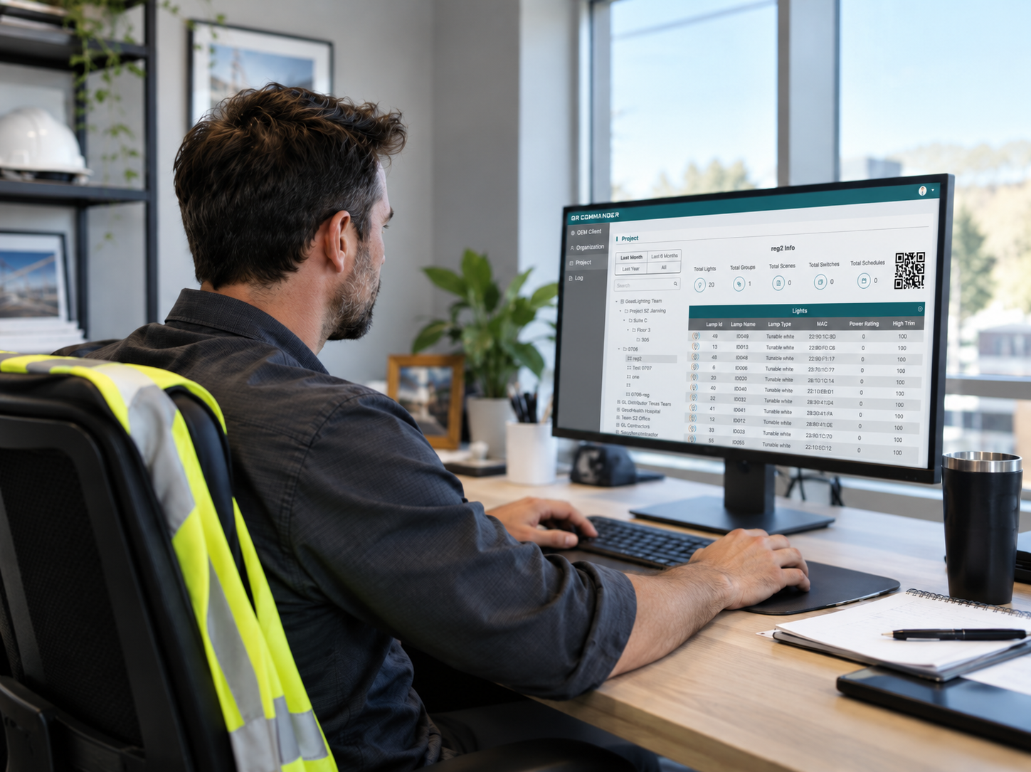 Project manager at a desk in a modern office reviewing the QR Commander lighting project management portal on a monitor